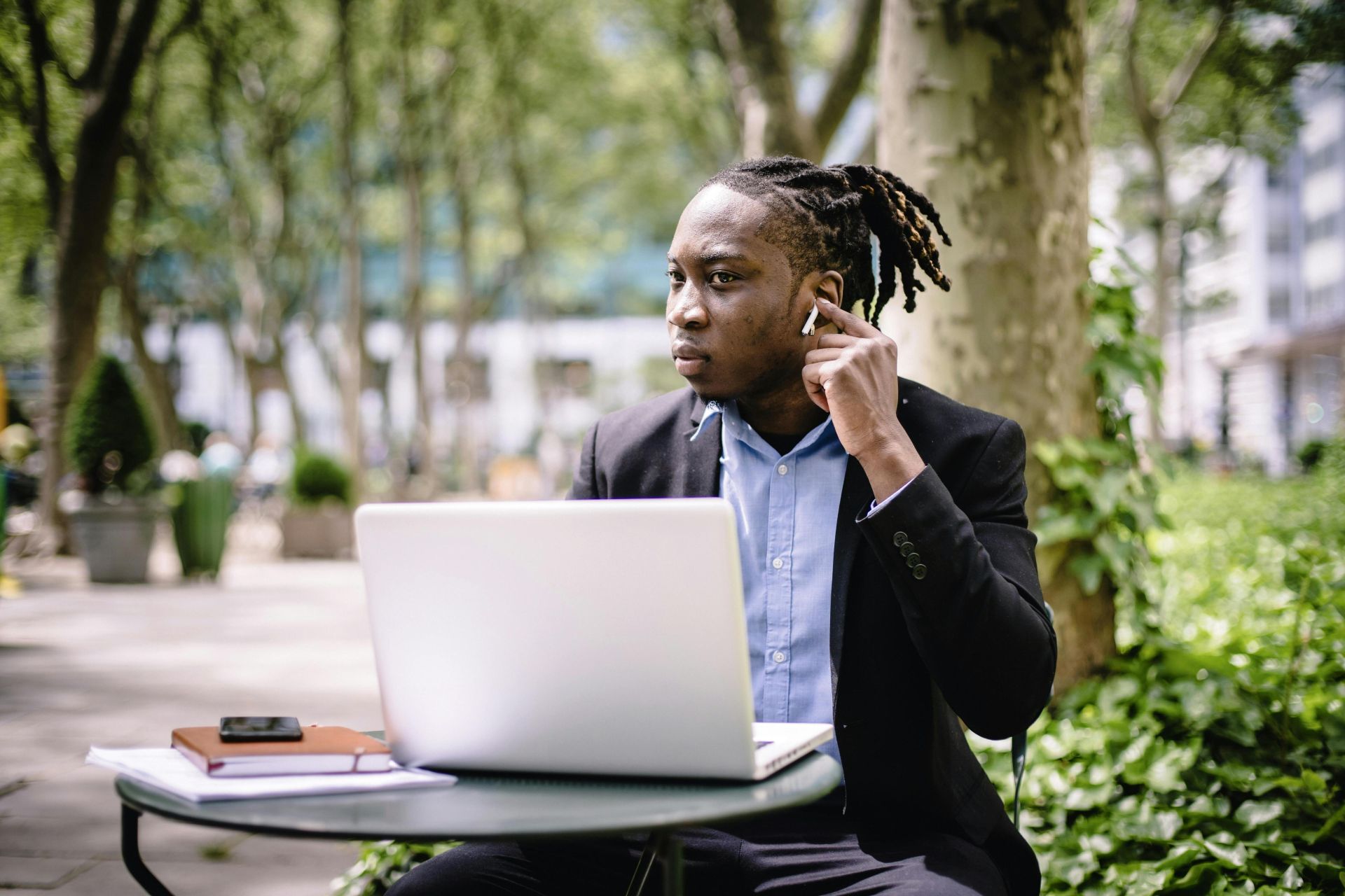 A businessman uses a laptop and earbuds while working outdoors in a sunny park.