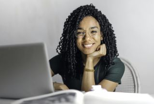 Joyful businesswoman with curly hair smiling at camera while using laptop indoors.